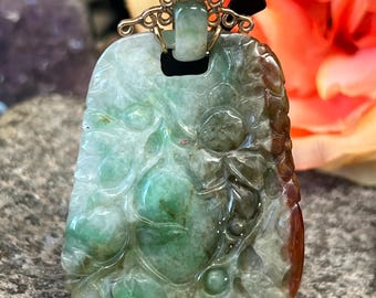 May include: A carved green jade pendant with a gold bail. The jade is a mottled green with brown and white accents. The pendant is shaped like a teardrop and has a floral design.