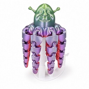 May include: A 3D printed articulated figure featuring a green head with trumpet-shaped ears and a smiling expression, connected to a series of dangling, segmented tentacles that transition in color from purple to red, supported by a white stand.