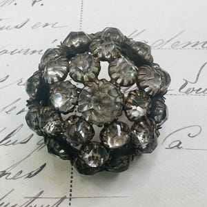 May include: A dome-shaped brooch featuring numerous clear, faceted glass stones set in a dark, aged metal frame. The stones are arranged in a cluster, creating a rounded, textured surface with a metallic base visible between the individual settings.