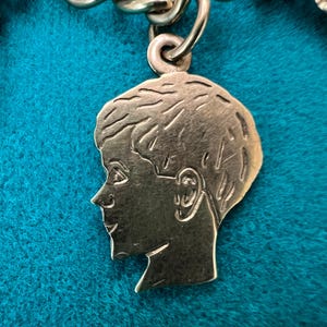 May include: A gold-toned charm in the shape of a person's head, with a side profile of a person with short hair. The charm is detailed with facial features and a textured surface.