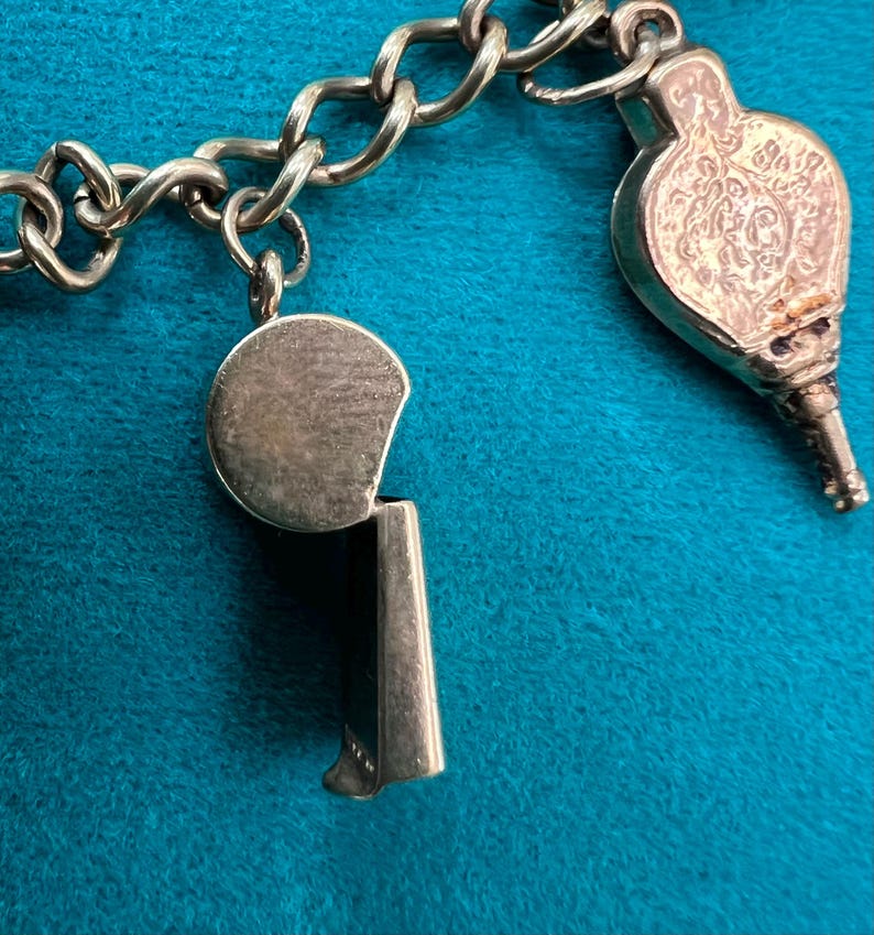 May include: A silver charm bracelet with a whistle charm and a decorative charm. The whistle charm is round with a silver finish. The decorative charm is ornate and has a silver finish.