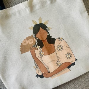May include: White tote bag featuring a stylized illustration of a woman in a traditional dress, holding a fan. The woman has dark hair and is adorned with floral accents. The bag has black handles.