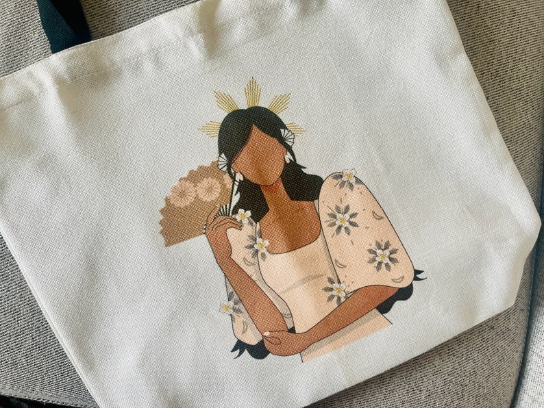 May include: White tote bag featuring a stylized illustration of a woman in a traditional dress, holding a fan. The woman has dark hair and is adorned with floral accents. The bag has black handles.