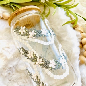 May include: A clear glass jar with a bamboo lid and a floral design. The design features white flowers and green leaves, resembling a lei. The jar is sitting on a white fluffy surface.