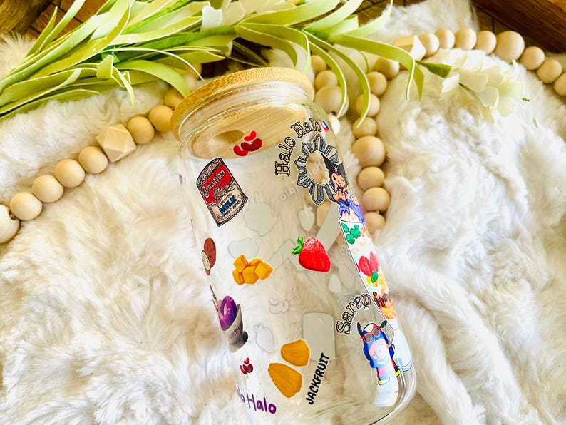 May include: A clear glass jar with a bamboo lid decorated with colorful stickers. The stickers feature images of fruit, food, and a cartoon character. The jar is labeled "Halo" and "Jackfruit".