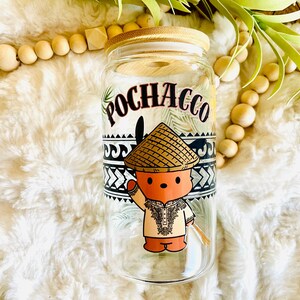 Puede incluir: Un vaso de vidrio transparente con tapa de bamb&uacute; con la palabra "POCHACCO" en escritura negra. El vaso tiene una ilustraci&oacute;n de dibujos animados de un personaje que lleva un sombrero c&oacute;nico y ropa tradicional. El dise&ntilde;o incluye patrones geom&eacute;tricos en blanco y negro.