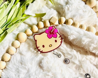 Puede incluir: Un personaje amarillo de Hello Kitty con una flor de hibisco rosa en la cabeza. El personaje está en un carrete de credencial de plástico transparente con un cordón retráctil plateado.