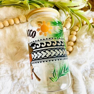 Puede incluir: Un vaso de vidrio transparente con tapa de bamb&uacute;, con un dise&ntilde;o colorido. El dise&ntilde;o incluye la palabra "Tacco", un sol, hojas de palma y patrones geom&eacute;tricos. El vaso est&aacute; decorado con una figura con sombrero de paja.