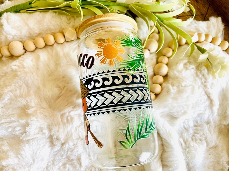 Puede incluir: Un vaso de vidrio transparente con tapa de bamb&uacute;, con un dise&ntilde;o colorido. El dise&ntilde;o incluye la palabra "Tacco", un sol, hojas de palma y patrones geom&eacute;tricos. El vaso est&aacute; decorado con una figura con sombrero de paja.