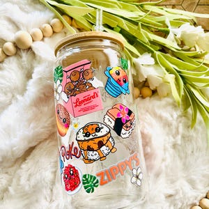 Puede incluir: Una botella de agua reutilizable de vidrio transparente con tapa de bamb&uacute; y una pajita. La botella est&aacute; decorada con pegatinas coloridas que representan comida y bebidas hawaianas, incluyendo poke, sushi, hielo raspado y donas de la panader&iacute;a Leonard's. Las pegatinas tienen caras de dibujos animados y el texto "Zippy's" y "Leonard's Bakery".