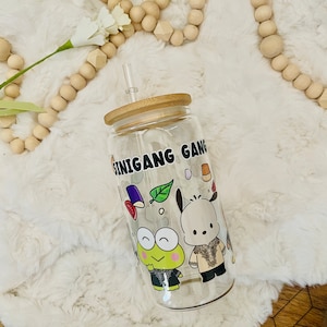 May include: Clear glass tumbler with a bamboo lid and straw. The tumbler features the text "SINIGANG GANG" and cartoon illustrations of characters and food items. The tumbler is on a white fluffy surface.