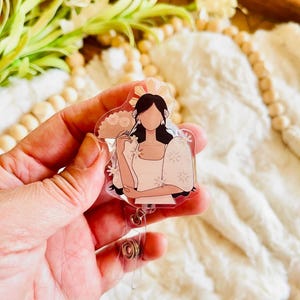 May include: A clear acrylic badge reel featuring a stylized illustration of a woman in a traditional Filipino dress. The design includes a sunburst headdress and floral details, with a retractable cord and clip.