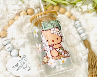 Puede incluir: Un vaso de vidrio transparente con tapa de bambú y una pajita. El vaso tiene un diseño de Hello Kitty con un collar de flores y hojas tropicales. Un cartel blanco en forma de corazón con el texto "You have whole h for wi" es visible.