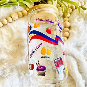 May include: A clear glass jar with a bamboo lid decorated with colorful stickers featuring the text "Halo Halo" and images of fruit, ice cream, and other ingredients. The jar is sitting on a white fluffy surface.