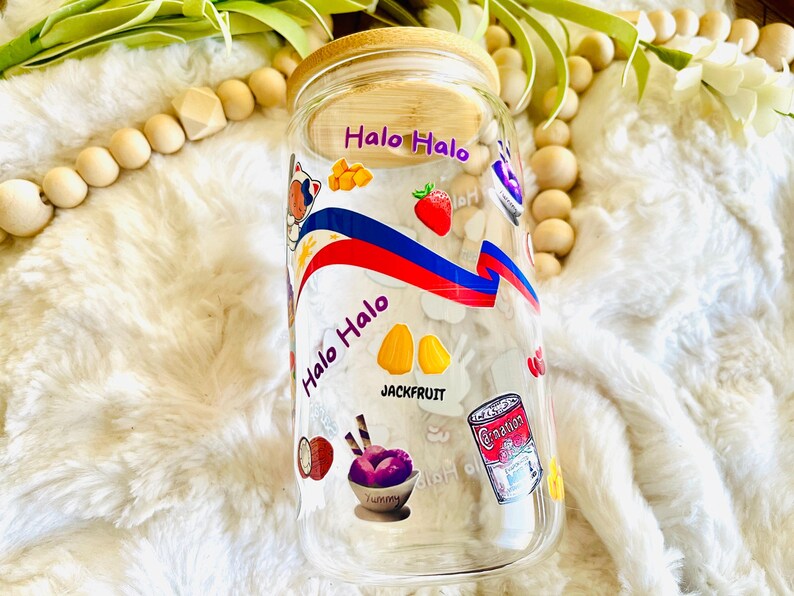 May include: A clear glass jar with a bamboo lid decorated with colorful stickers featuring the text "Halo Halo" and images of fruit, ice cream, and other ingredients. The jar is sitting on a white fluffy surface.