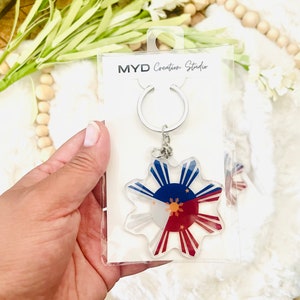 May include: A silver keyring with a clear acrylic charm featuring the Philippine flag. The charm is shaped like a sun with eight rays. The flag is red, white, and blue with a yellow sun and three stars in the centre. The keyring is packaged in a clear plastic bag with a white card that reads "MYD Creation Studio".