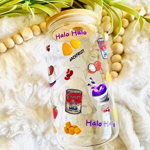 May include: A clear glass jar with a wooden lid and colorful stickers featuring the words "Halo Halo" and images of various ingredients, including jackfruit, ube, and condensed milk.