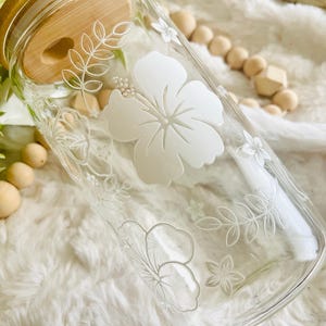 May include: A clear glass jar with a bamboo lid and white etched hibiscus flowers and leaves. The jar is sitting on a white fuzzy surface.