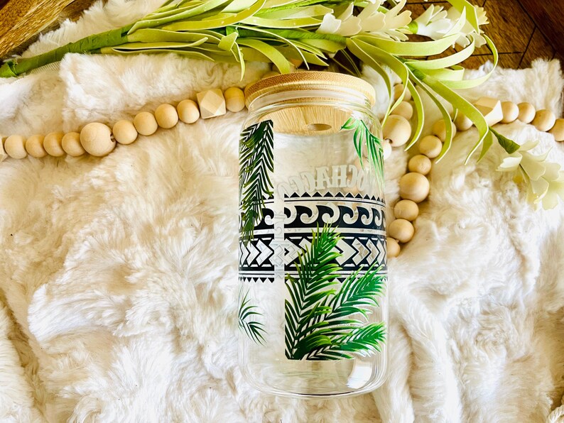 Puede incluir: Un vaso de vidrio transparente con tapa de bamb&uacute;, con un dise&ntilde;o tropical con hojas de palmera y patrones geom&eacute;tricos en blanco y negro. La palabra "MAHALO" est&aacute; impresa en el vaso. El vaso est&aacute; rodeado de elementos decorativos.