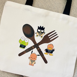 May include: A white canvas tote bag with a brown wooden spoon, fork, and a green frog, a black bird, a brown duck, and a brown bear wearing black jackets. The spoon and fork are crossed in the middle of the bag.