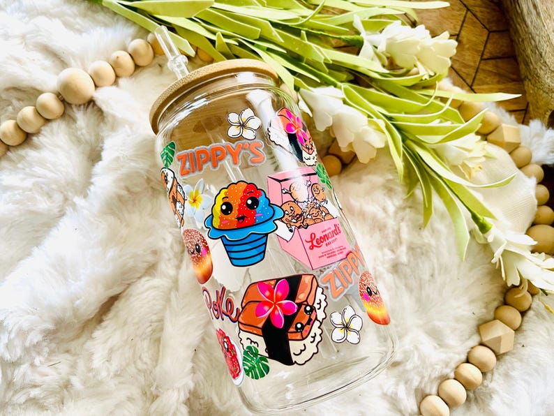 Puede incluir: Una botella de agua reutilizable de vidrio transparente con tapa de bamb&uacute; y una pajita. La botella est&aacute; decorada con pegatinas coloridas que representan comida y flores hawaianas. Las pegatinas incluyen el texto "Zippy's" y "Leonard's Bakery".
