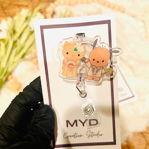 May include: A clear acrylic badge holder with a cartoon image of two characters. The characters are a yellow cat with a pink bow and a brown bear wearing a blue and white patterned shirt. The badge holder has a retractable cord and the text "MYD Creation Studio" is printed on the bottom.