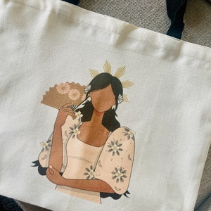 May include: White tote bag featuring a stylized illustration of a woman in a traditional outfit, holding a fan. The artwork includes floral details and a gold halo. Black handles complete the design.