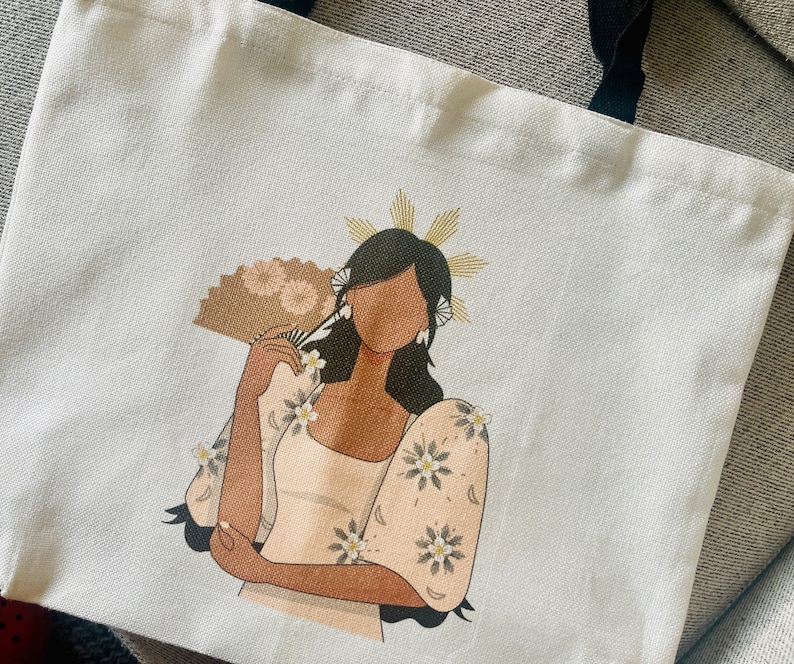 May include: White tote bag featuring a stylized illustration of a woman in a traditional outfit, holding a fan. The artwork includes floral details and a gold halo. Black handles complete the design.