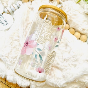 May include: A clear glass jar with a wooden lid, decorated with pink flowers and gold swirls. The jar is sitting on a white fluffy surface.