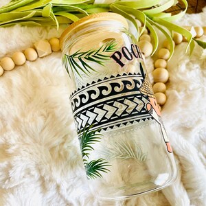 Puede incluir: Vaso de vidrio transparente con tapa de bamb&uacute;, con un dise&ntilde;o de inspiraci&oacute;n polinesia. El dise&ntilde;o incluye patrones geom&eacute;tricos en blanco y negro, hojas de palma y la palabra "Poco" en rosa. El vaso descansa sobre una superficie blanca y esponjosa.