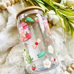 May include: A clear glass reusable water bottle with a bamboo lid and a straw. The bottle is decorated with colorful stickers of fruit, flowers, and a frog.