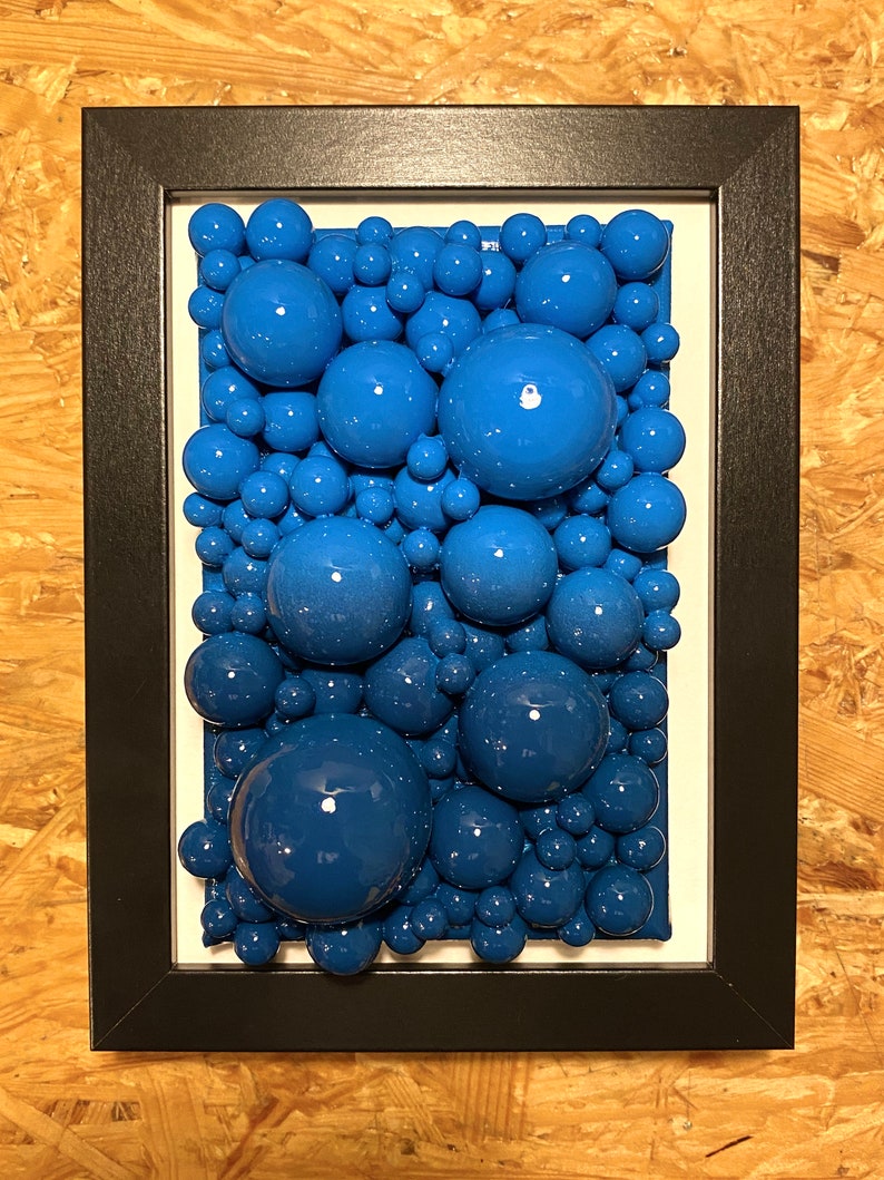 May include: A framed artwork featuring a cluster of glossy, blue spheres of varying sizes. The spheres are arranged densely against a white background, creating a textured, three-dimensional effect. The frame is black.