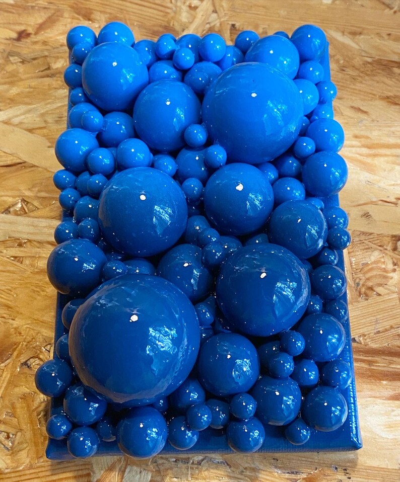 May include: A rectangular art piece featuring a vibrant arrangement of glossy, deep blue spheres of varying sizes. The spheres are densely packed, creating a textured, three-dimensional effect. The artwork is set against a dark blue background.