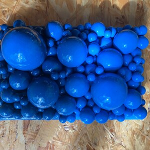 May include: A rectangular abstract art piece featuring a cluster of glossy, blue spheres of varying sizes. The spheres are densely packed together, creating a textured surface. The artwork is mounted on a wooden panel.