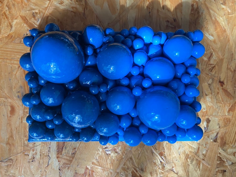 May include: A rectangular abstract art piece featuring a cluster of glossy, blue spheres of varying sizes. The spheres are densely packed together, creating a textured surface. The artwork is mounted on a wooden panel.