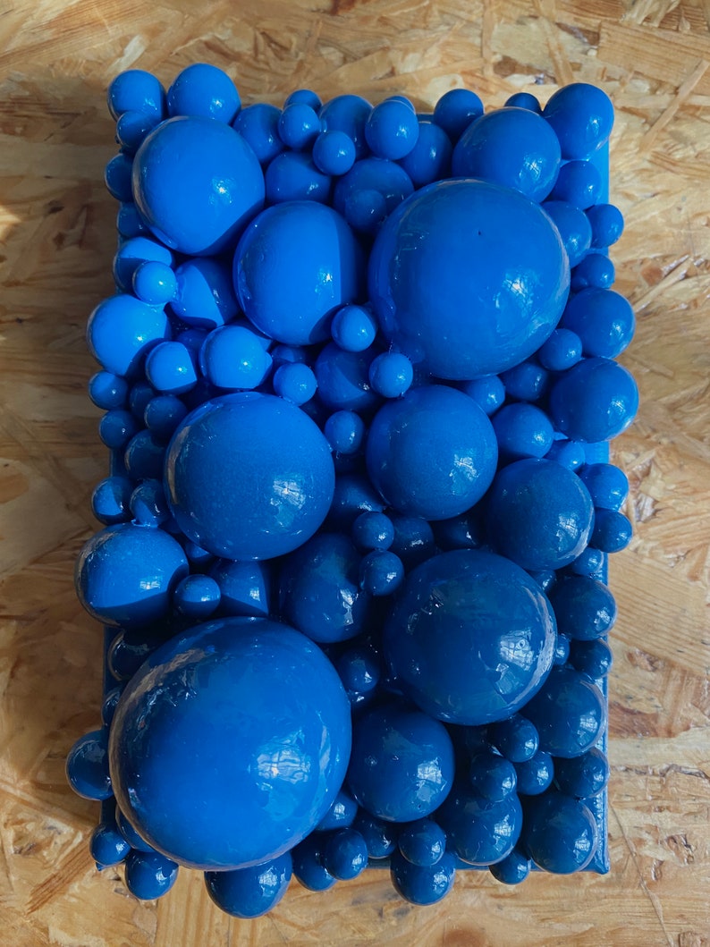 May include: A rectangular wall art piece featuring a collection of glossy, blue spheres of varying sizes. The spheres are arranged densely, creating a textured, three-dimensional effect. The artwork is painted in shades of blue.