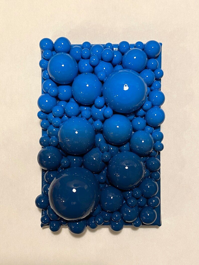 May include: Abstract art piece featuring a rectangular frame filled with glossy, blue spheres of varying sizes. The spheres create a textured, three-dimensional effect, with a gradient of shades from light to dark blue.