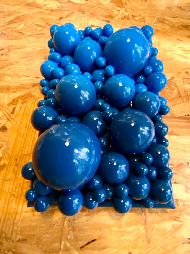 May include: A rectangular sculpture composed of various sizes of glossy, deep blue spheres. The spheres are clustered together, creating a textured, three-dimensional effect. The sculpture is set against a light brown, wood-grain background.