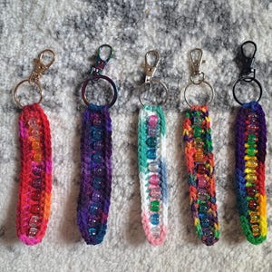 May include: Five handmade keychains with colorful crochet straps and clear beads. The keychains feature a variety of colors, including red, purple, green, and yellow. Each keychain has a metal clasp and ring for attaching keys or other items.