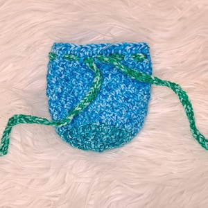 May include: A small, handmade crochet pouch in shades of blue and turquoise, with a drawstring closure. The drawstring is a contrasting green and white. The pouch is likely used for small items or as a gift bag.