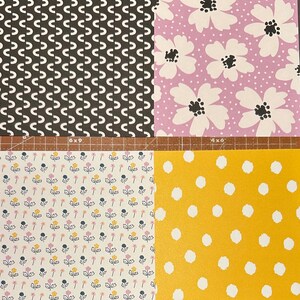 May include: Four different patterned fabric swatches. The first swatch is black with a white repeating pattern of curved lines. The second swatch is light purple with a white repeating pattern of flowers. The third swatch is white with a repeating pattern of small pink, yellow, and blue flowers. The fourth swatch is yellow with a repeating pattern of white circles.