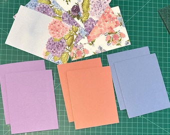 May include: A collection of stationery items including several rectangular cards featuring watercolor-style floral patterns with pink, purple, and blue hydrangea blossoms, alongside solid-colored cardstock in lavender, coral, and periwinkle blue.