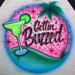 May include: A pink and blue airbrushed design on a white t-shirt featuring a margarita glass with a lime wedge, a palm tree, and the text "Gettin' Buzzed".
