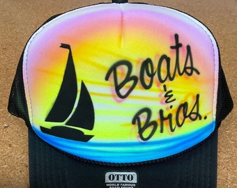 May include: A black and blue trucker hat with a sunset and sailboat design. The hat has the words "Boats & Bros." written in black cursive on the front.