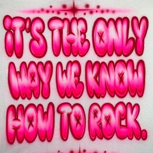 May include: Pink and white airbrushed design on a white t-shirt. The design says "It's the only way we know how to rock."