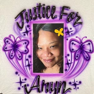 May include: A white t-shirt with a purple and black airbrushed design. The design features a portrait of a woman, two butterflies, and the words "Justice For Arynn".