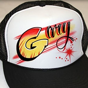 Op de afbeelding: Een zwart-witte truckercap met een wit voorpaneel. Het voorpaneel heeft een rood, geel en oranje airbrush-ontwerp met de naam "Gary" met een stervormige uitbarsting.