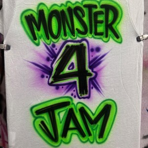 Op de afbeelding: Een wit T-shirt met een groen, zwart en paars airbrush-ontwerp. Het ontwerp bevat de woorden "Monster 4 Jam" in graffiti-stijl.