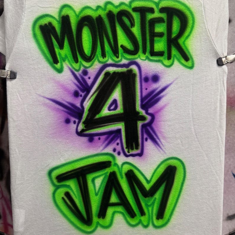 Op de afbeelding: Een wit T-shirt met een groen, zwart en paars airbrush-ontwerp. Het ontwerp bevat de woorden "Monster 4 Jam" in graffiti-stijl.