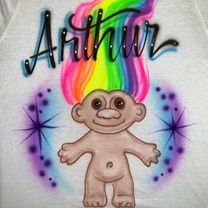 May include: A white t-shirt with a rainbow-colored troll with the name "Arthur" written in black above it. The troll has brown eyes and a brown nose. The background is white with purple and blue stars.